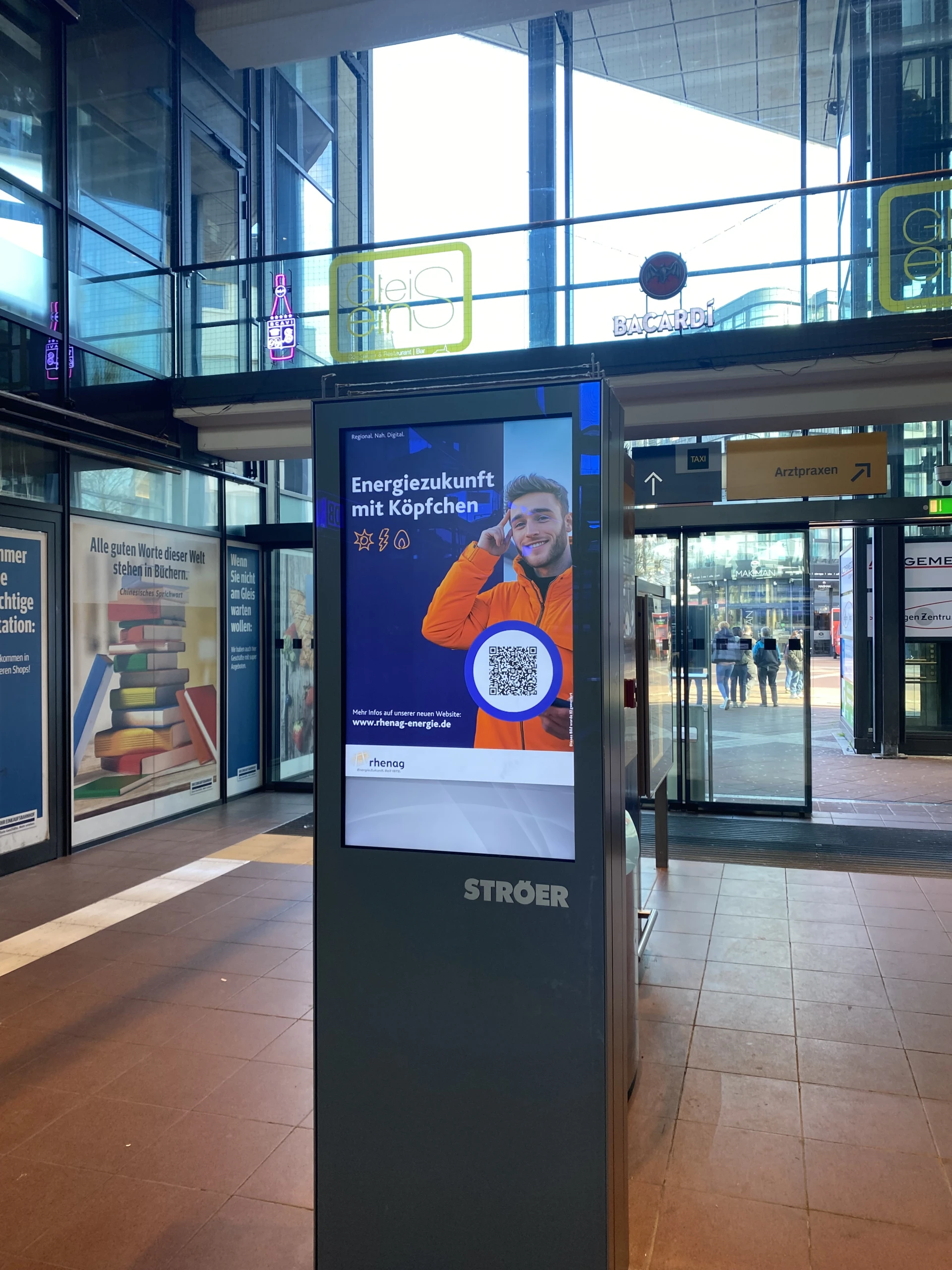 rhenag Kampagne für rhenag-energie.de mit "Energiezukunft mit Köpfchen" als Headline und einem KI-generierten Bild, Icons und QR-Code auf digitaler Werbefläche in einem Bahnhof